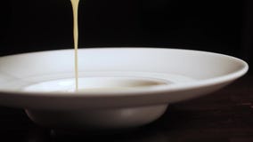 Pouring cream soup from a metal pan into white plate with wide fields. Close-up