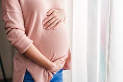 Pregnant woman touching belly while standing near window at home stock photos Pregnant woman touching belly while standing near window at home stock photos