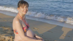 Pregnant young woman in a swimsuit sitting on the beach near the sea