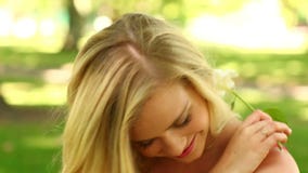 Pretty blonde relaxing in the park smiling at the camera while smelling a flower