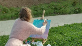 A pretty woman with red hair, paints a picture on canvas, which stands on the easel. The lady is in the open air near stock video footage A pretty woman with red hair, paints a picture on canvas, which stands on the easel. The lady is in the open air near stock video footage