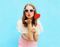 Pretty woman in sunglasses with red heart lollipop sends an air kiss over colorful blue Royalty Free Stock Photography Pretty woman in sunglasses with red heart lollipop sends an air kiss over colorful blue Royalty Free Stock Photography