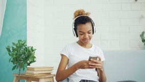 Pretty young woman student is texting friends chatting online and listening to music through headphones resting on sofa