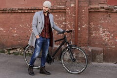 Przystojny m?ody cz?owiek pcha jego roweru puszek w popielatym ?akiecie i kapeluszu ulica w mie?cie zdjęcie stock Przystojny m?ody cz?owiek pcha jego roweru puszek w popielatym ?akiecie i kapeluszu ulica w mie?cie zdjęcie stock