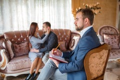 Psykolog med en anteckningsbok på en stol, i bakgrunden ett par på en soffa som ser de royaltyfri foto Psykolog med en anteckningsbok på en stol, i bakgrunden ett par på en soffa som ser de royaltyfri foto