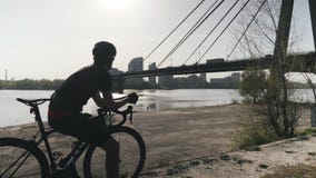 Radfahrer in der schwarzen Sportkleidung auf dem schwarzen Fahrrad, das in Richtung des Flusses und der Br?cke tr?umend ?ber Zuku Radfahrer in der schwarzen Sportkleidung auf dem schwarzen Fahrrad, das in Richtung des Flusses und der Br?cke tr?umend ?ber Zuku
