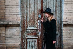 Ragazza bionda con capelli lunghi, in cappotto nero in cappello, supporti sui precedenti di vecchia casa con mattoni a vista dell immagine stock Ragazza bionda con capelli lunghi, in cappotto nero in cappello, supporti sui precedenti di vecchia casa con mattoni a vista dell immagine stock