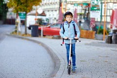 Ragazzo attivo del bambino della scuola nella guida del casco di sicurezza con il suo motorino nella città con lo zaino il giorno immagini stock Ragazzo attivo del bambino della scuola nella guida del casco di sicurezza con il suo motorino nella città con lo zaino il giorno immagini stock