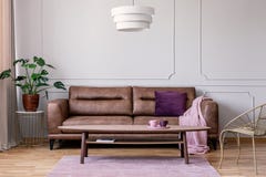 Real photo of brown leather sofa with violet cushion and pastel pink blanket standing in light grey sitting room interior with Mon stock photo Real photo of brown leather sofa with violet cushion and pastel pink blanket standing in light grey sitting room interior with Mon stock photo
