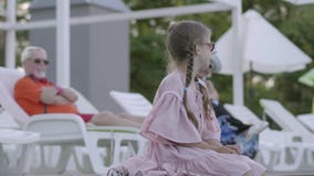 Reife Paare, die auf sunbeds nahe dem Pool auf dem Hintergrund liegen Wenig lustiges Mädchen mit den Zöpfen, die am Rand sitzen stock video footage Reife Paare, die auf sunbeds nahe dem Pool auf dem Hintergrund liegen Wenig lustiges Mädchen mit den Zöpfen, die am Rand sitzen stock video footage