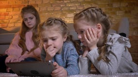 Retrato de três meninas caucasianos pequenas que encontram-se no assoalho e que olham na tabuleta atentamente na atmosfera de cas filme Retrato de três meninas caucasianos pequenas que encontram-se no assoalho e que olham na tabuleta atentamente na atmosfera de cas filme