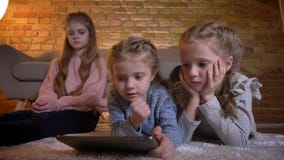 Retrato de três meninas caucasianos pequenas que encontram-se no assoalho e que olham na tabuleta seriamente na atmosfera de casa filme Retrato de três meninas caucasianos pequenas que encontram-se no assoalho e que olham na tabuleta seriamente na atmosfera de casa filme