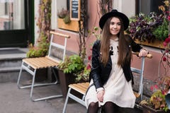 Retrato de una muchacha feliz sonriente del inconformista de los jóvenes que se sienta en una silla de madera cerca del edificio Foto de archivo Retrato de una muchacha feliz sonriente del inconformista de los jóvenes que se sienta en una silla de madera cerca del edificio Foto de archivo