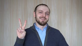 Retrato del hombre barbudo joven que gesticula la muestra de la victoria, dentro almacen de video Retrato del hombre barbudo joven que gesticula la muestra de la victoria, dentro almacen de video