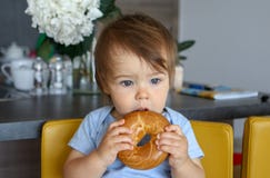 Retrato do bebê pensativo bonito com o corte de cabelo à moda que guarda e que come o bagel grande com boca aberta foto de stock Retrato do bebê pensativo bonito com o corte de cabelo à moda que guarda e que come o bagel grande com boca aberta foto de stock