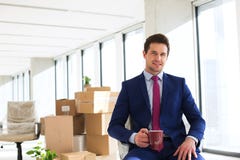 Retrato do homem de negócios novo que come o café com as caixas moventes no fundo no escritório Fotografia de Stock Retrato do homem de negócios novo que come o café com as caixas moventes no fundo no escritório Fotografia de Stock