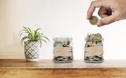 Räcka att sätta myntet i glasflaskor, med garneringväxten på träskrivbordet, sparande pengar, investering och hushålla begrepp Royaltyfri Bild Räcka att sätta myntet i glasflaskor, med garneringväxten på träskrivbordet, sparande pengar, investering och hushålla begrepp Royaltyfri Bild