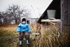 Sad little boy in a blue jacket and brown pants sitting on the bench. Next to the wooden structure. Gloomy day in the village. Sad little boy in a blue jacket royalty free stock photography Sad little boy in a blue jacket and brown pants sitting on the bench. Next to the wooden structure. Gloomy day in the village. Sad little boy in a blue jacket royalty free stock photography