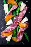 Salada com o serrano do jamon do presunto, camembert, melão, rúcula na placa de pedra preta da ardósia no fundo preto Vista super fotografia de stock Salada com o serrano do jamon do presunto, camembert, melão, rúcula na placa de pedra preta da ardósia no fundo preto Vista super fotografia de stock