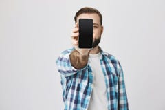 Samiec model z brodą i modny ostrzyżenie w sprawdzać koszulowym pokazuje smartphone przy kamery nakrycia twarzą z nim, stojący zdjęcie royalty free Samiec model z brodą i modny ostrzyżenie w sprawdzać koszulowym pokazuje smartphone przy kamery nakrycia twarzą z nim, stojący zdjęcie royalty free