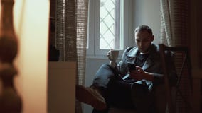 Sammanträde för ung man i fåtölj i vardagsrum och användasmartphone Attraktiv manlig bläddrande internet, drinkkaffe lager videofilmer Sammanträde för ung man i fåtölj i vardagsrum och användasmartphone Attraktiv manlig bläddrande internet, drinkkaffe lager videofilmer