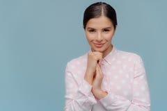Satisfied confident lovely female with smile on face, keeps hands near chin, looks at camera, has dark combed hair, wears polka stock photography Satisfied confident lovely female with smile on face, keeps hands near chin, looks at camera, has dark combed hair, wears polka stock photography