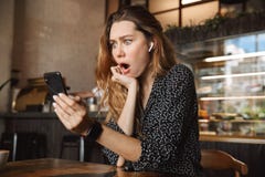 Scared shocked displeased beautiful young pretty woman sitting in cafe indoors using mobile phone talking with friends stock photo Scared shocked displeased beautiful young pretty woman sitting in cafe indoors using mobile phone talking with friends stock photo
