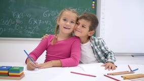 Schulliebe, Mitschüler schreiben während der Lektion bei Tisch und betrachten dann Kamera und Lächeln auf Hintergrund der Tafel stock footage Schulliebe, Mitschüler schreiben während der Lektion bei Tisch und betrachten dann Kamera und Lächeln auf Hintergrund der Tafel stock footage