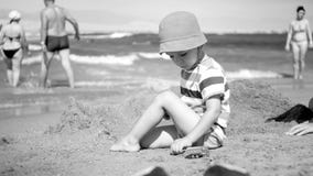 Schwarzweiss-Bild des netten Kleinkindjungen im Hut, der auf dem Seestrand sitzt und mit Spielzeugautos spielt Entspannendes Kind lizenzfreie stockbilder Schwarzweiss-Bild des netten Kleinkindjungen im Hut, der auf dem Seestrand sitzt und mit Spielzeugautos spielt Entspannendes Kind lizenzfreie stockbilder