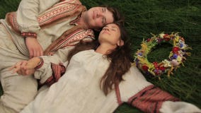 Schönes Brunettemädchen im traditionellen ukrainischen Hemd mit Blumenkranz mit ihrem Freund, der am grünen Feld liegt stock footage Schönes Brunettemädchen im traditionellen ukrainischen Hemd mit Blumenkranz mit ihrem Freund, der am grünen Feld liegt stock footage