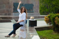 Schönes Brunettemädchen sitzt auf der Straße mit einem Laptop und einem Einkauf Sie bewegt ihre Hand wellenartig stockfoto Schönes Brunettemädchen sitzt auf der Straße mit einem Laptop und einem Einkauf Sie bewegt ihre Hand wellenartig stockfoto
