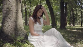 Schönes junges Mädchen des Porträts mit dem langen brunette Haar, das ein langes weißes Sommermodekleid herein sitzt unter einem stock video Schönes junges Mädchen des Porträts mit dem langen brunette Haar, das ein langes weißes Sommermodekleid herein sitzt unter einem stock video