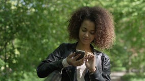 Schönes junges Mädchen mit dem dunklen gelockten Haar unter Verwendung ihres Handys, im Freien Schönes junges Mädchen mit dem dunklen gelockten Haar unter Verwendung ihres Handys, im Freien