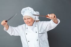 Senior woman chef studio standing isolated on gray holding knives scary smiling playful close-up stock photo Senior woman chef studio standing isolated on gray holding knives scary smiling playful close-up stock photo