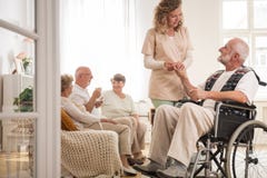 Senior man on wheelchair with helpful nurse holding his hand and friends sitting on couch drinking tea stock photography Senior man on wheelchair with helpful nurse holding his hand and friends sitting on couch drinking tea stock photography