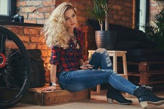 Sensual blonde hipster girl with long curly hair dressed in a fleece shirt and jeans holds a smartphone sitting on a. Wooden box at a studio with loft interior Royalty Free Stock Photos Sensual blonde hipster girl with long curly hair dressed in a fleece shirt and jeans holds a smartphone sitting on a. Wooden box at a studio with loft interior Royalty Free Stock Photos