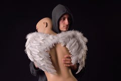 Serious man dressed in black hoodie holding in his arms a plasti. C woman body mannequin with white feathered wings behind the back on dark background. Lack of Stock Photography Serious man dressed in black hoodie holding in his arms a plasti. C woman body mannequin with white feathered wings behind the back on dark background. Lack of Stock Photography