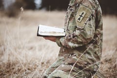Shallow focus shot of a young soldier kneeling while holding an open bible in a field