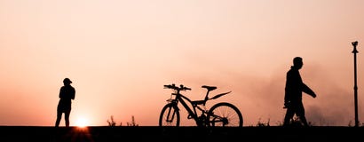 Silhouette du fonctionnement de l'homme et de femme, faisant du vélo et marchant sur le passage couvert à la rive dans la soirée Photo libre de droits Silhouette du fonctionnement de l'homme et de femme, faisant du vélo et marchant sur le passage couvert à la rive dans la soirée Photo libre de droits