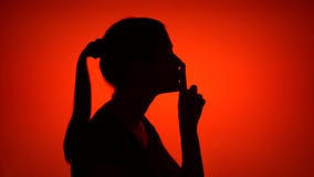 Silhouette of young woman making silence gesture on red background. Concept of mystery and secrecy