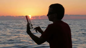 Silhouettieren Sie Mann mit digitaler Tablette in den Händen am Sonnenuntergangstrand Die Sonne wird fast hinter den Ozean einges stock video footage Silhouettieren Sie Mann mit digitaler Tablette in den Händen am Sonnenuntergangstrand Die Sonne wird fast hinter den Ozean einges stock video footage