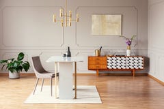 Silla en la tabla debajo de la lámpara del oro en interior de la sala de estar con el cartel sobre armario con la planta Foto ver foto de archivo Silla en la tabla debajo de la lámpara del oro en interior de la sala de estar con el cartel sobre armario con la planta Foto ver foto de archivo