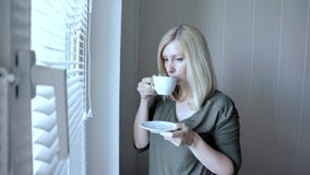 Situación rubia hermosa thinkful triste joven de la mujer cerca de la ventana con las persianas por la mañana y el café de consum almacen de video Situación rubia hermosa thinkful triste joven de la mujer cerca de la ventana con las persianas por la mañana y el café de consum almacen de video