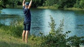 A slender gray-haired man in a blue t-shirt, cap and glasses walks barefoot on the green grass on the river Bank on a stock footage A slender gray-haired man in a blue t-shirt, cap and glasses walks barefoot on the green grass on the river Bank on a stock footage