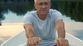 A slender gray-haired serious man in a gray t-shirt and glasses rowing on a white boat on a calm river on a summer stock video A slender gray-haired serious man in a gray t-shirt and glasses rowing on a white boat on a calm river on a summer stock video