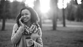 Smartphone de fala da menina atrativa da raça misturada e café bebendo no parque da cidade com sacos Jovem mulher após a compra s fotografia de stock Smartphone de fala da menina atrativa da raça misturada e café bebendo no parque da cidade com sacos Jovem mulher após a compra s fotografia de stock