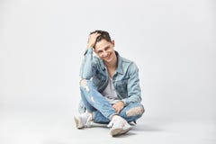Smiling dark-haired guy dressed in a white t-shirt, jeans and a denim jacket is sitting cross-legged on the floor on the stock image Smiling dark-haired guy dressed in a white t-shirt, jeans and a denim jacket is sitting cross-legged on the floor on the stock image