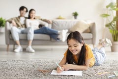 Smiling japanese youngsters girl study at home, writing, drawing in notebook and millennial parents sits and rest. On sofa in living room interior. Covid-19