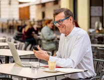 Smiling old man checking smart phone while working on computer in terrace outdoors coffee shop in seniors using modern technology royalty free stock images Smiling old man checking smart phone while working on computer in terrace outdoors coffee shop in seniors using modern technology royalty free stock images