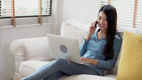 Sofá que se sienta asiático hermoso de la mujer joven usando el ordenador portátil y el teléfono elegante móvil que habla en el d Sofá que se sienta asiático hermoso de la mujer joven usando el ordenador portátil y el teléfono elegante móvil que habla en el d
